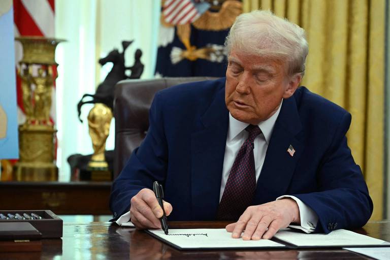 Um homem está sentado em uma mesa， escrevendo com uma caneta em um documento. Ele usa um terno escuro e uma gravata. Ao fundo， há uma bandeira dos Estados Unidos e uma decoração de escritório com um busto e uma pintura. O ambiente parece ser um escritório formal.