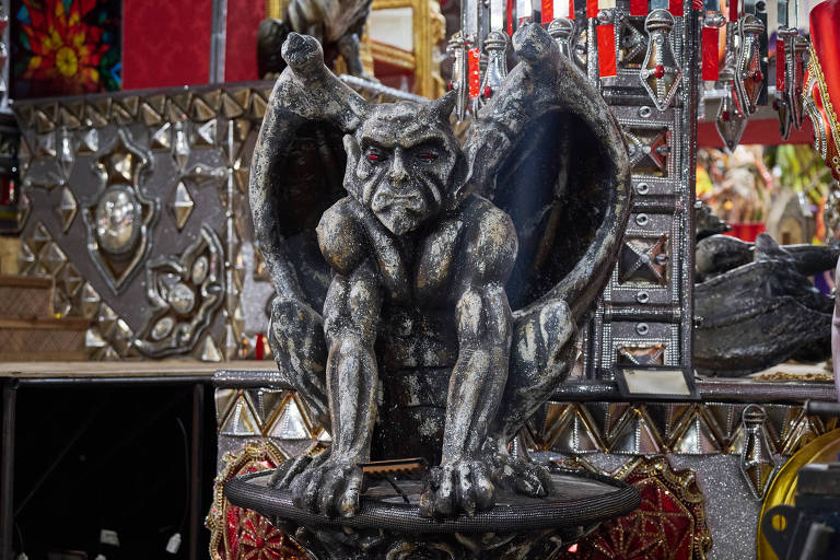 A imagem mostra uma estátua de um demônio com asas, sentado em uma base decorativa. O demônio tem uma expressão facial intensa, com olhos vermelhos e músculos bem definidos. O fundo apresenta elementos decorativos metálicos e coloridos, sugerindo um ambiente festivo ou de celebração.