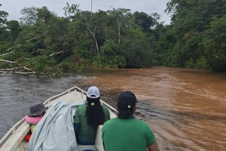 A imagem mostra a vista de duas pessoas em uma pequena embarcação navegando em um rio na floresta amazônica. A água do rio é de cor marrom e há vegetação densa ao redor, com árvores e folhagens. As pessoas estão usando roupas de proteção e chapéus, e uma delas está coberta com um tecido. O ambiente parece úmido e tropical.