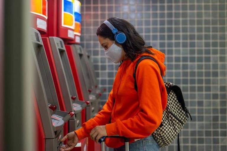 Uma jovem mulher está utilizando um caixa eletrônico em um ambiente interno. Ela está vestindo um moletom laranja e fones de ouvido azuis， além de uma máscara facial. A mulher está concentrada na tela do caixa eletrônico， que é vermelho， e há um fundo de azulejos cinzas.