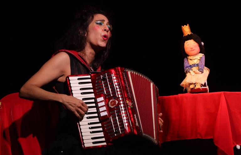 Uma artista está tocando um acordeão vermelho enquanto canta. Ela tem cabelo longo e escuro， e usa maquiagem colorida. À sua frente， há uma mesa vermelha com uma boneca sentada， que usa um vestido amarelo e uma coroa. O fundo é escuro， destacando a performance.