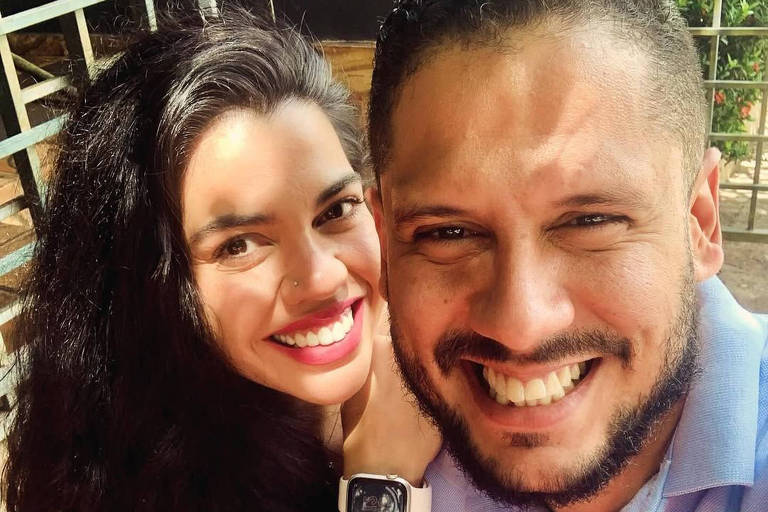 Uma selfie de um casal sorridente. A mulher tem cabelo longo e escuro， usa uma blusa com estampas florais em tons de rosa e branco. O homem tem cabelo curto e barba， veste uma camisa azul clara. Ambos estão em um ambiente iluminado， com uma grade ao fundo.