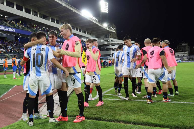 Um grupo de jogadores de futebol está se cumprimentando e celebrando após uma partida. Eles estão vestindo uniformes brancos e azuis， com alguns usando coletes rosa. O cenário é um estádio iluminado à noite， com torcedores ao fundo.