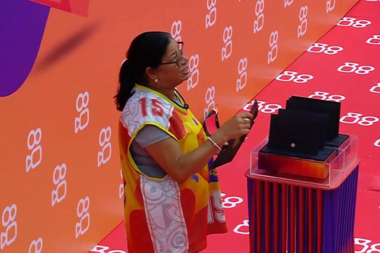 Uma mulher está em pé ao lado de um pedestal com um dispositivo eletrônico em cima. Ela usa uma camiseta colorida com o número 15 e está segurando um objeto na mão. O fundo é laranja com o logotipo '88' repetido em branco.
