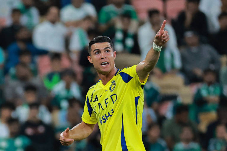 Cristiano Ronaldo， vestindo um uniforme amarelo， aponta o dedo indicador da mão esquerda para o alto， com torcedores vestindo verde e branco ao fundo