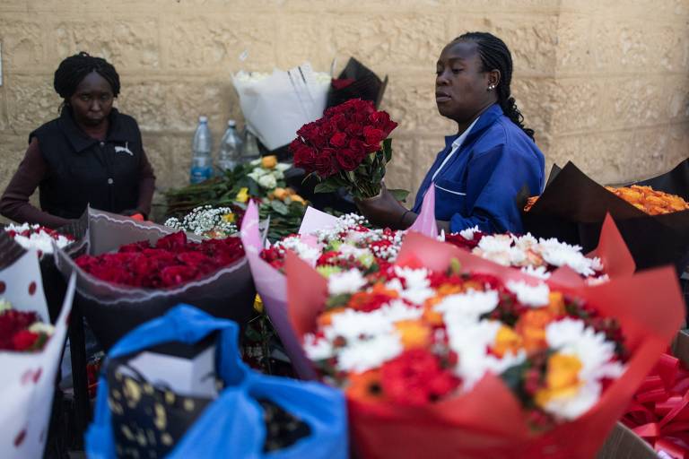 A imagem mostra duas mulheres em um mercado de flores. A mulher à direita está segurando um buquê de rosas vermelhas e parece estar organizando as flores. A mulher à esquerda está em segundo plano, observando. Ao redor delas, há diversos arranjos de flores em papéis coloridos, incluindo rosas vermelhas, brancas e flores de outras cores. O fundo é uma parede de pedra clara.