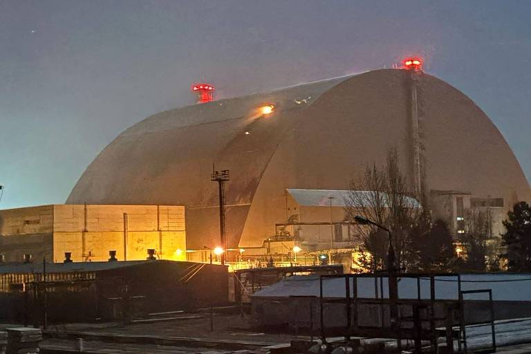 A imagem mostra a estrutura do sarcófago que cobre o reator nuclear de Chernobyl， iluminada à noite. A construção é grande e arredondada， com luzes vermelhas visíveis no topo. O ambiente ao redor parece ser uma área industrial， com algumas estruturas metálicas e um piso de concreto visível em primeiro plano.