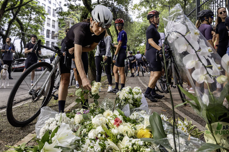 A imagem mostra um grupo de ciclistas em uma homenagem. Um ciclista， vestindo roupas escuras e um capacete branco， está se agachando para colocar uma rosa branca em um monte de flores que inclui rosas brancas e amarelas， além de outras flores. Ao fundo， outros ciclistas estão parados， alguns em bicicletas， e há árvores e prédios ao redor.