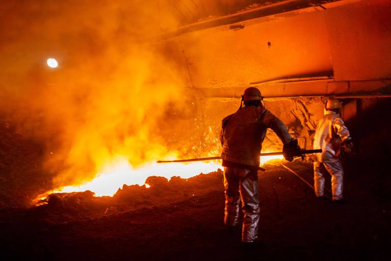 A imagem mostra dois trabalhadores em um ambiente industrial， provavelmente em uma fundição. Eles estão usando roupas de proteção prateadas e capacetes， enquanto manipulam material incandescente. O fundo é iluminado por uma intensa luz laranja e amarela， proveniente de um forno， criando uma atmosfera de calor e atividade intensa.