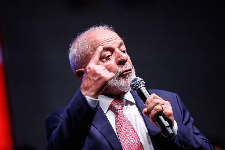 Um homem de cabelo grisalho e barba， vestido com um terno escuro e uma gravata vermelha， está falando ao microfone. Ele levanta um dedo， possivelmente enfatizando um ponto em seu discurso， enquanto se dirige a uma audiência.