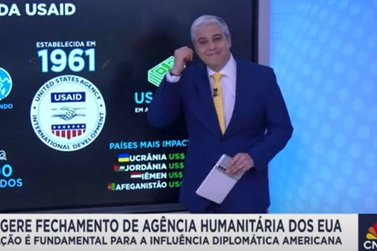 Um apresentador de notícias está em pé, sorrindo e fazendo um gesto com a mão, enquanto segura um bloco de notas. Ele está vestido com um terno azul e uma gravata amarela. Ao fundo, há um gráfico que menciona a USAID, estabelecida em 1961, e lista países impactados como Ucrânia, Jordânia, Iémen e Afeganistão, com valores em dólares. A parte inferior da imagem contém um texto sobre o fechamento da agência humanitária dos EUA.
