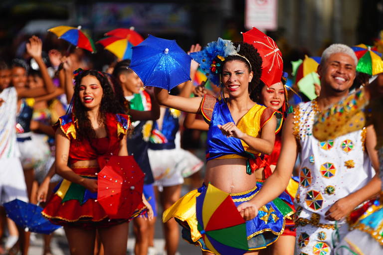 Um grupo de pessoas participa de um desfile de Carnaval， vestindo trajes coloridos. Algumas pessoas seguram guarda-chuvas azuis e vermelhos， enquanto dançam e sorriem. O ambiente é festivo， com muitas cores vibrantes e uma atmosfera alegre.