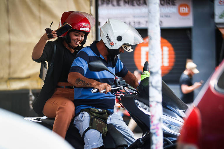 A imagem mostra um homem pilotando uma motocicleta com uma mulher como passageira. Ambos estão usando capacetes， sendo que a mulher está com um capacete vermelho e o homem com um capacete branco. Ela usa uma blusa preta e calça bege， e ele veste uma camisa polo com listras azuis e pretas horizontais. Eles estão em um ambiente urbano， com outros veículos e pessoas ao fundo.