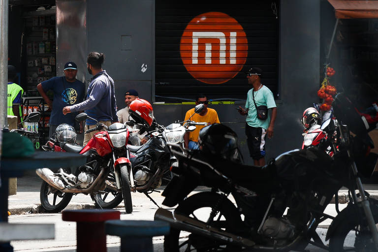 A imagem mostra um cenário urbano com várias motocicletas estacionadas. No fundo, há uma loja com o logotipo da marca 'mi' em um círculo laranja pintado num portão de ferro fechado. Algumas pessoas estão conversando em pé, enquanto outras estão sentadas. O ambiente parece movimentado, com a presença de mais motocicletas e pessoas ao redor.