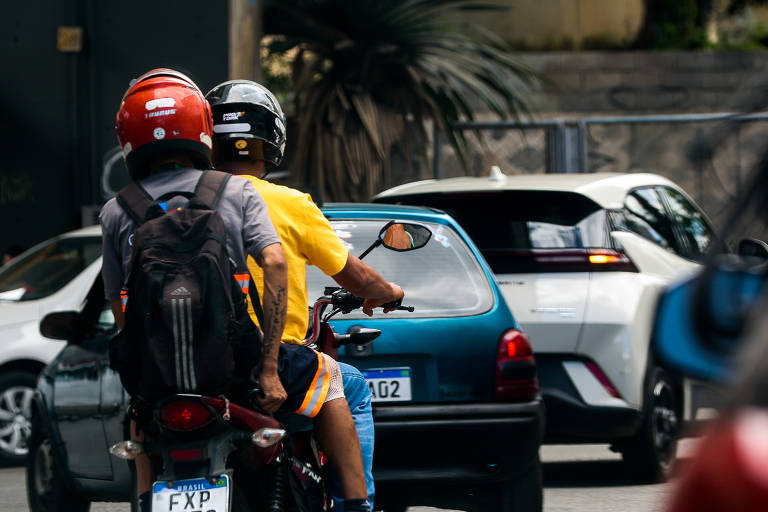 A imagem mostra dois motociclistas em uma rua movimentada. O motociclista da frente está usando um capacete preto e uma camiseta amarela， enquanto o passageiro atrás usa um capacete vermelho e uma mochila. Ao fundo， há um carro azul e outros veículos parados no trânsito.