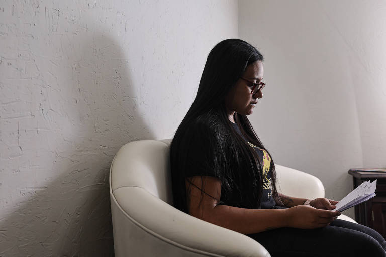 A imagem mostra uma mulher sentada em um sofá branco， lendo documentos. Ela tem cabelo longo e liso， e usa óculos escuros. O ambiente é iluminado， com uma parede clara ao fundo e uma mesa ao lado.