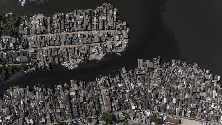 Imagem aérea mostrando uma área urbana densa, com várias construções de telhados cinzentos. A área é cercada por água, com um rio ou canal visível. As edificações estão dispostas de forma irregular, criando um padrão caótico. A vegetação é escassa, e a imagem apresenta uma paleta de cores predominantemente cinza e verde.