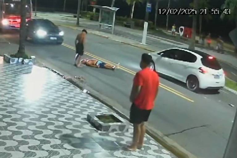 A imagem mostra uma cena capturada por uma câmera de segurança em uma rua. Há um homem em pé， vestindo uma camiseta vermelha， observando uma pessoa caída no chão. Um carro branco está parado próximo à pessoa caída， enquanto outro veículo se aproxima. O pavimento é de ladrilhos e a iluminação é noturna.