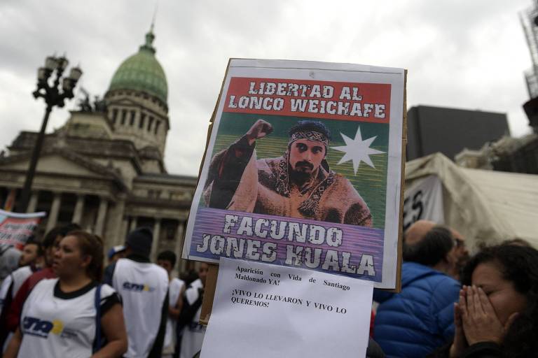 A imagem mostra um cartaz em um protesto， com a frase 039;Libertad al Longo Weichafe039; e a imagem de um homem com cabelo longo e barba. O fundo apresenta o edifício do Congresso Nacional da Argentina， com pessoas ao redor. O cartaz também menciona 039;Facundo Jones Huala039; e 039;Mapuche039; em referência à sua identidade. O clima é de manifestação， com pessoas vestindo camisetas brancas.