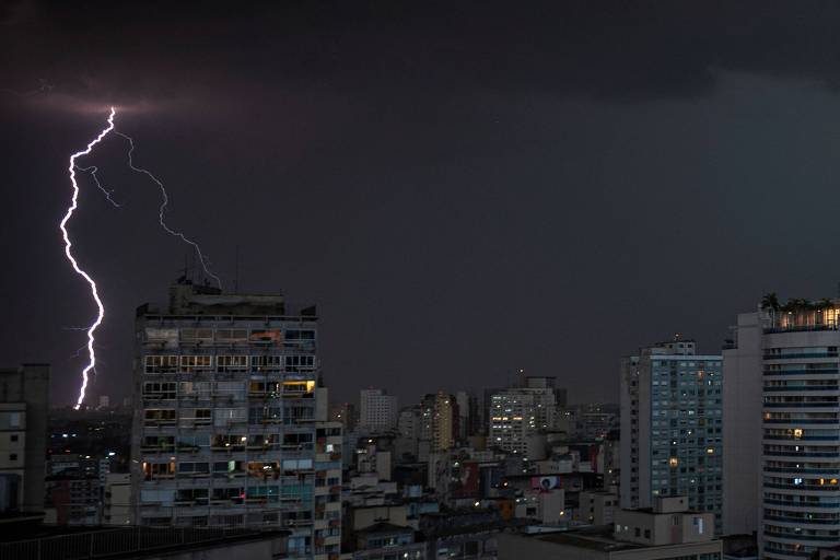 A imagem mostra um céu escuro com nuvens carregadas e um raio iluminando a cena. Várias edificações urbanas estão visíveis， com janelas iluminadas e uma variedade de alturas e estilos arquitetônicos. O ambiente transmite uma sensação de tempestade iminente.