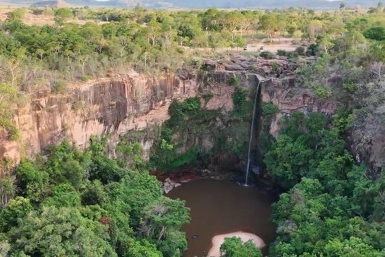 A imagem mostra uma vista aérea de uma cachoeira que deságua em um lago cercado por vegetação densa. As paredes ao redor da cachoeira são formadas por rochas e a água cai de uma altura considerável. O ambiente é predominantemente verde, com árvores e arbustos ao redor, e ao fundo, é possível ver uma área mais clara, possivelmente uma estrada ou campo.