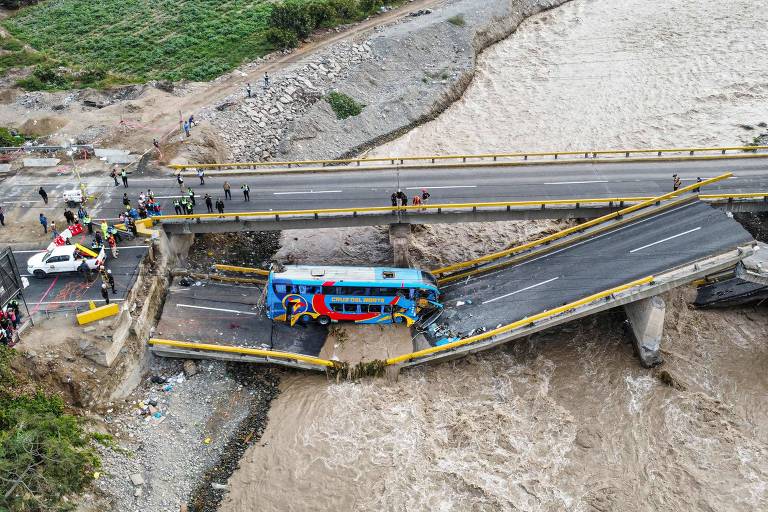 A imagem mostra uma ponte danificada， com uma parte dela colapsada e inclinada. Um ônibus colorido está parcialmente na ponte， enquanto a água de um rio em cheia flui abaixo. Há várias pessoas e veículos ao redor， incluindo carros de emergência. O cenário é de destruição， com vegetação ao fundo.