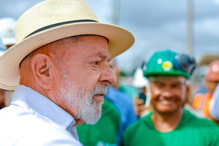 A imagem mostra um homem de perfil， usando um chapéu de palha e uma camisa branca. Ele tem uma barba grisalha e parece estar em um evento ao ar livre. Ao fundo， há pessoas vestindo camisetas verdes e capacetes de segurança， sorrindo e observando. O céu está parcialmente nublado.