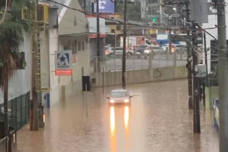 A imagem mostra uma rua inundada， com água cobrindo a maior parte da via. Um carro está parcialmente submerso， com as luzes traseiras acesas， emitindo um brilho laranja. Ao fundo， é possível ver edifícios e postes de luz， além de sinais de trânsito. A cena reflete uma situação de alagamento em uma área urbana.