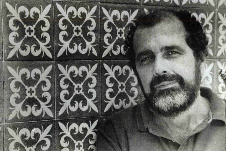 A imagem mostra um homem com barba e cabelo escuro， posando em frente a um fundo de azulejos com padrões florais em preto e branco. Ele está usando uma camisa de gola e tem uma expressão serena.