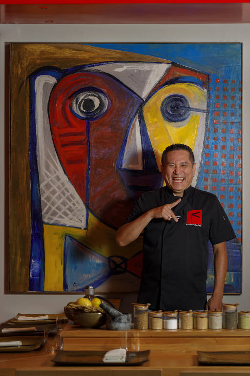 Um chef está posando em frente a uma grande pintura colorida que apresenta formas abstratas e rostos. Ele está usando um uniforme preto com um logotipo vermelho e sorri enquanto aponta para a câmera. Na mesa à sua frente, há vários recipientes com especiarias e utensílios de cozinha.