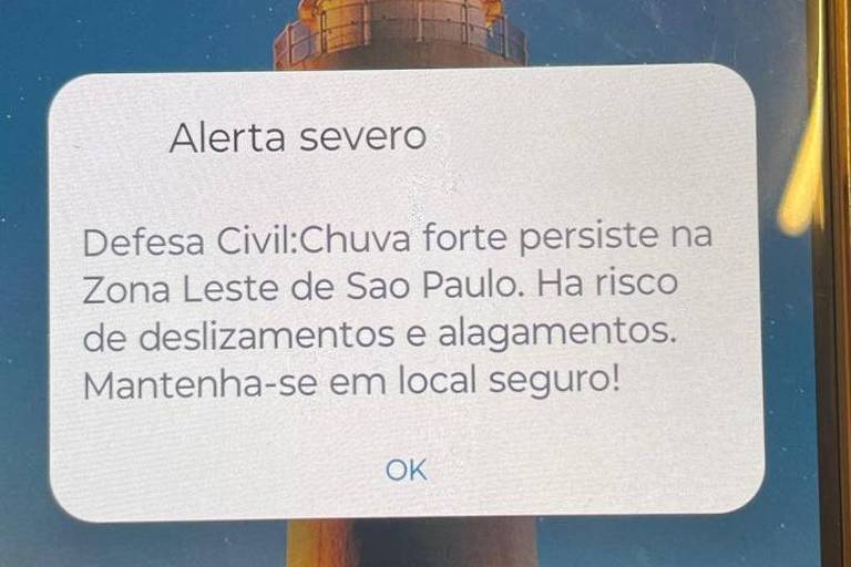 A imagem mostra um alerta severo na tela de um dispositivo， informando sobre a persistência de chuva forte na Zona Leste de São Paulo. O texto alerta sobre o risco de deslizamentos e alagamentos， recomendando que as pessoas se mantenham em local seguro.