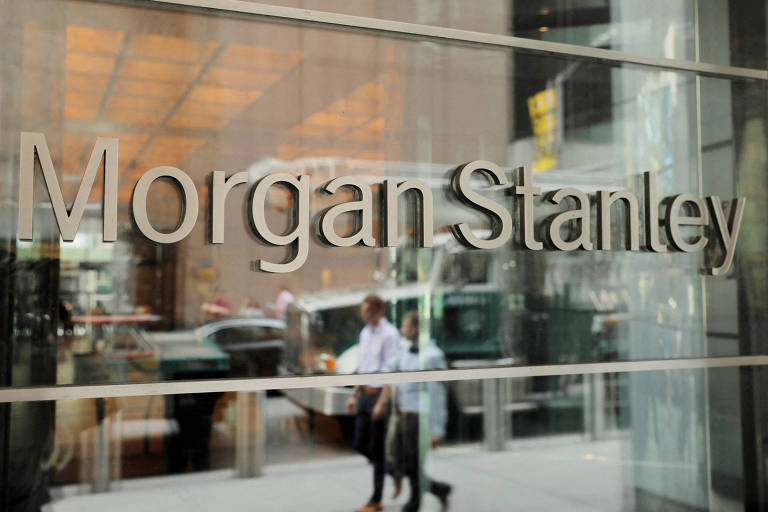 A imagem mostra a fachada de um escritório da Morgan Stanley， com o nome da empresa em letras grandes e prateadas. Ao fundo， é possível ver pessoas caminhando e veículos na rua， além de uma iluminação interna visível através das janelas.