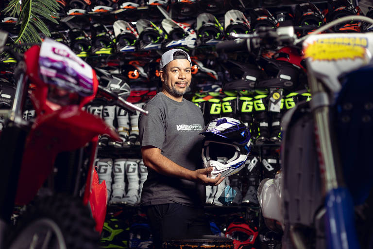 Um homem está em uma loja de equipamentos de motocross， segurando um capacete. Ele usa uma camiseta cinza e um boné. Ao fundo， há uma prateleira cheia de capacetes e botas de motocross， com cores vibrantes como verde e preto. Também são visíveis algumas motos de motocross na parte frontal da imagem.