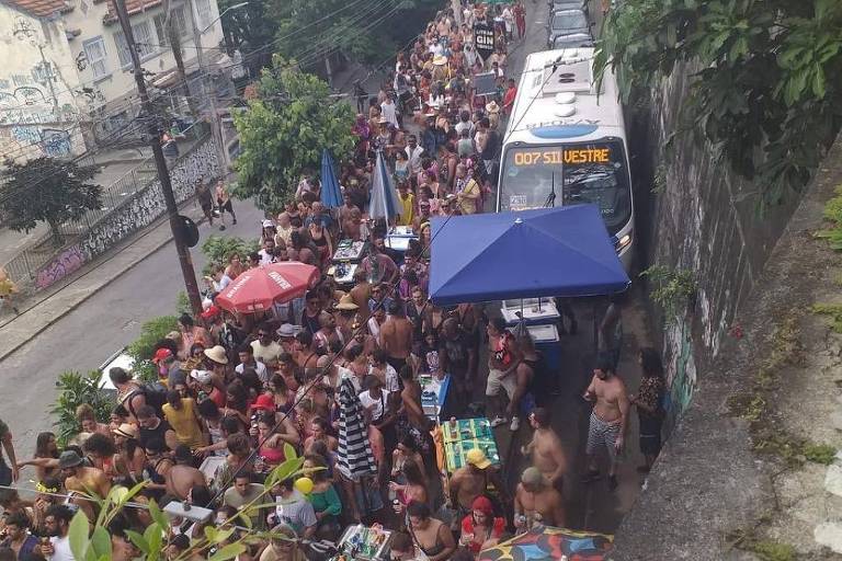 A imagem mostra uma grande multidão reunida em uma festa de rua， com pessoas sem camisa e algumas usando chapéus. Há barracas com toldos coloridos， incluindo um em azul e outro em vermelho. Ao fundo， é possível ver um ônibus e vegetação ao redor. A cena é animada， com muitas pessoas interagindo entre si.