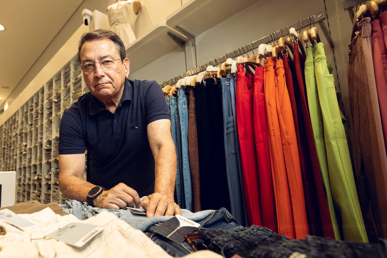 Um homem de meia-idade está em uma loja de roupas， olhando para a câmera enquanto se inclina sobre uma mesa com roupas. Ele usa uma camisa polo preta e tem óculos. Ao fundo， há uma parede com várias calças de cores diferentes， incluindo azul， vermelho e verde. A loja é bem iluminada e organizada.