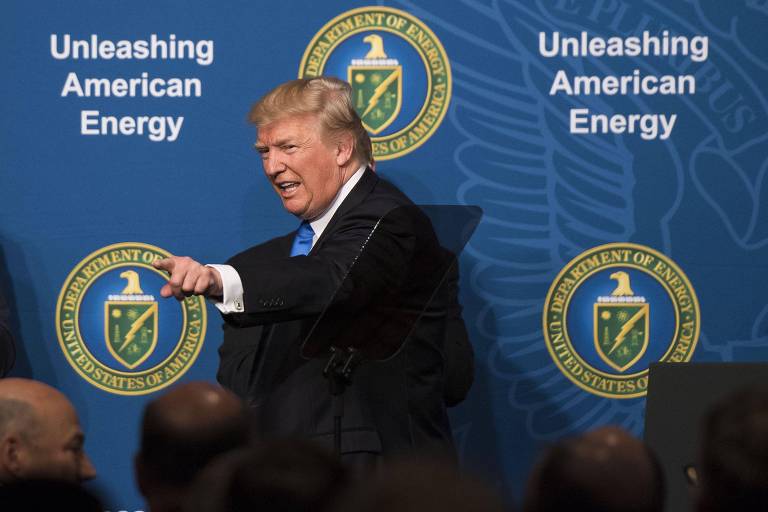 Um homem em um paletó escuro e gravata azul está em pé， gesticulando com a mão direita enquanto fala em um evento. Ao fundo， há um painel azul com o texto 039;Unleashing American Energy039; e emblemas do Departamento de Energia dos Estados Unidos.