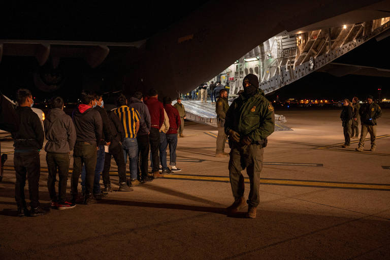 A imagem mostra um grupo de pessoas em fila， aguardando para embarcar em uma aeronave. A cena ocorre à noite， com iluminação artificial visível. Um agente de segurança， vestido com uniforme e máscara， está em pé próximo à fila， observando. A parte traseira da aeronave está aberta， permitindo acesso ao interior.