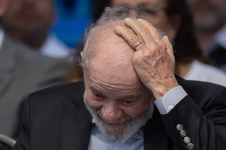 Imagem mostra o presidente Lula， um homem idoso， com cabelo grisalho e barba. Ele passa uma das mãos na cabeça， demonstrando preocupação. Ele usa um terno escuro e uma camisa clara. 