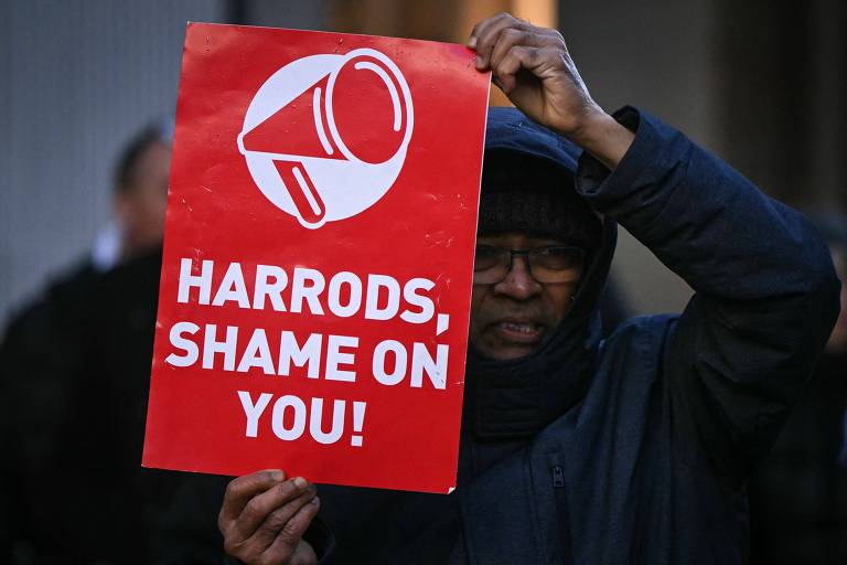 Uma pessoa segurando um cartaz vermelho com um megafone e as palavras 039;HARRODS， SHAME ON YOU!039; em letras brancas. A pessoa está vestida com um casaco escuro e um gorro， e parece estar participando de um protesto.