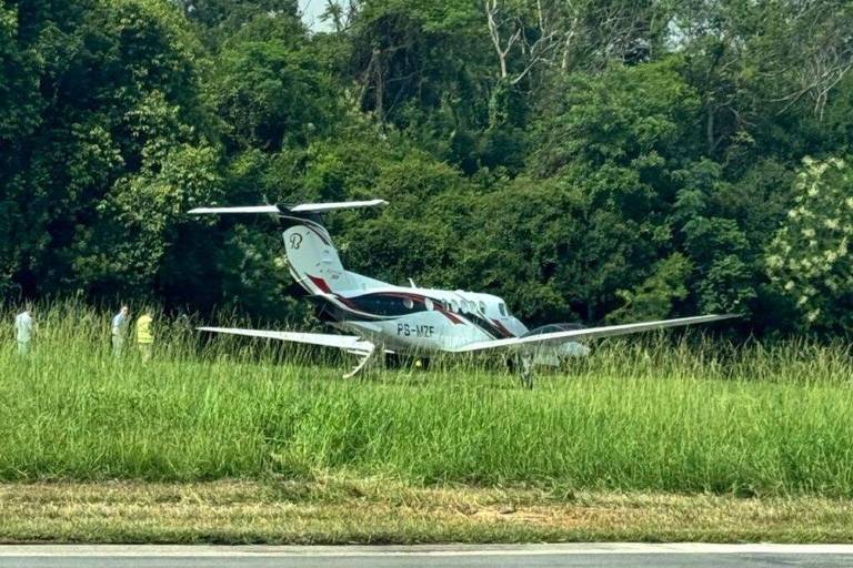 Um pequeno avião está pousado em uma área gramada， cercado por vegetação densa. Ao fundo， há árvores e arbustos. Algumas pessoas vestindo coletes amarelos estão próximas ao avião， e a pista de pouso é visível na parte inferior da imagem.