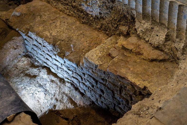 A imagem mostra uma área de escavação arqueológica com paredes de tijolos expostas. O solo é irregular e apresenta uma iluminação suave que destaca a textura dos tijolos e do terreno ao redor. Ao fundo， há uma estrutura com colunas， parcialmente visível， que sugere a presença de uma construção antiga.