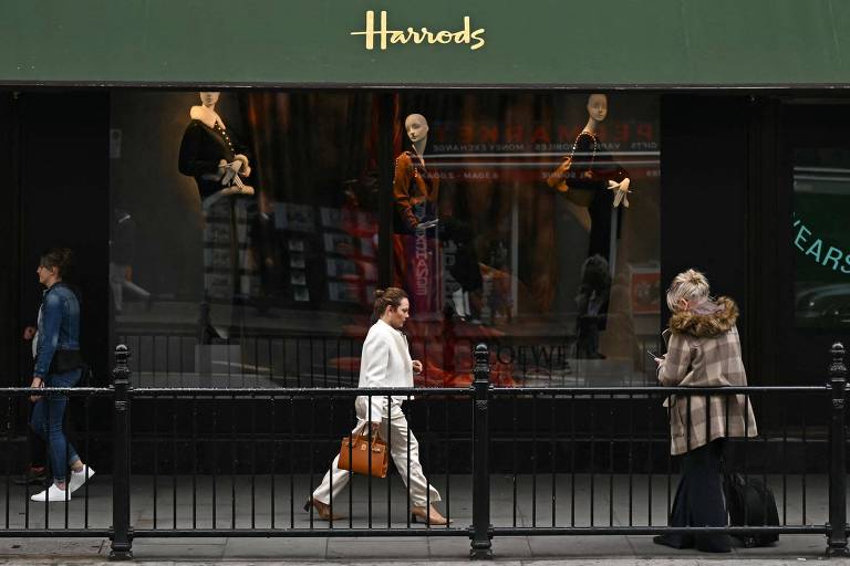 A imagem mostra a vitrine da loja Harrods， com manequins exibindo roupas em um fundo escuro. Na calçada， três pessoas estão caminhando: uma mulher vestindo um conjunto branco e carregando uma bolsa laranja， um homem com uma camiseta e calça jeans， e uma mulher com um casaco de pele e saia longa. O nome 039;Harrods039; está visível na parte superior da vitrine.