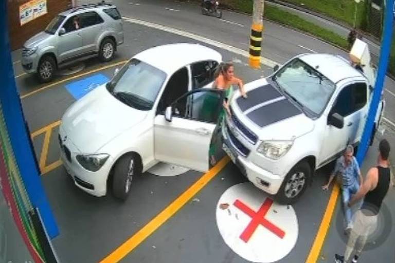 A imagem mostra um incidente em um estacionamento， onde um carro branco está parcialmente estacionado com a porta aberta e uma mulher saindo dele. Um veículo SUV de cor clara está estacionado ao lado， enquanto um homem está próximo ao carro branco. Ao fundo， outro carro pode ser visto， e há uma marca de 039;X039; no chão， indicando uma área restrita.