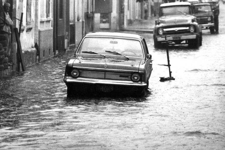 A imagem em preto e branco mostra uma rua inundada， com um carro parado em meio à água. O veículo está parcialmente submerso， e ao fundo， outro carro pode ser visto. À esquerda， uma pessoa está em pé， observando a cena. O ambiente é urbano， com casas visíveis ao longo da rua.