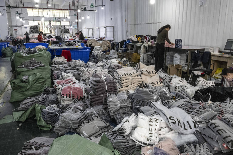 A imagem mostra um armazém com pilhas de roupas organizadas em sacos. Há várias pessoas trabalhando no local， algumas estão em mesas， enquanto outras estão em pé. O ambiente é amplo， com paredes brancas e iluminação fluorescente. Os sacos de roupas são predominantemente em tons de cinza， preto e bege， com algumas etiquetas visíveis com a palavra 039;SHEIN039;.