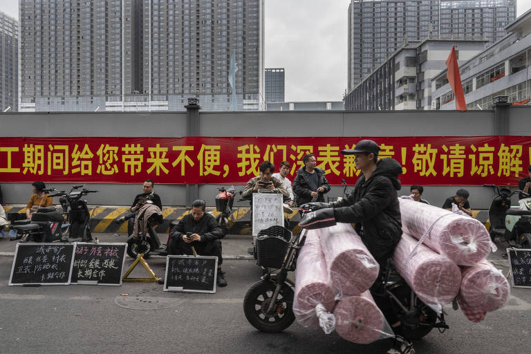 A imagem mostra um protesto em uma rua， onde várias pessoas estão sentadas em cadeiras， algumas segurando placas com textos. Ao fundo， há uma grande faixa vermelha com texto em chinês. Um motociclista passa pela cena， transportando rolos de material， possivelmente plástico ou papel， na parte traseira da moto. Edifícios altos são visíveis ao fundo.