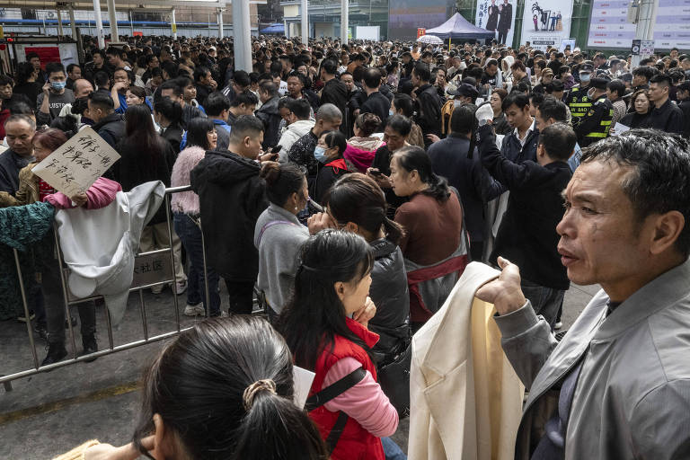 A imagem mostra uma grande multidão de pessoas em um espaço coberto， possivelmente aguardando para entrar em um evento. Algumas pessoas estão segurando cartazes， enquanto outras conversam entre si. A atmosfera parece ser de expectativa， com muitos indivíduos visíveis em diferentes direções. O ambiente é urbano， com estruturas metálicas e banners ao fundo.