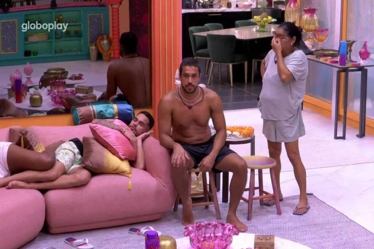 A imagem mostra um ambiente interno com um sofá grande e colorido. Três pessoas estão presentes: um homem sem camisa sentado no sofá, outro homem deitado com as pernas sobre o sofá e uma mulher em pé ao lado, com uma expressão pensativa. O ambiente é decorado com objetos coloridos e há uma mesa ao fundo. A iluminação é suave e acolhedora.