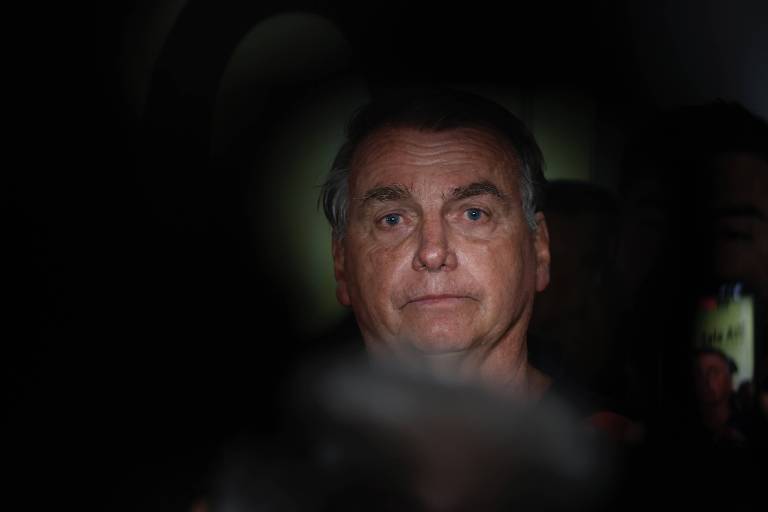 A imagem mostra Bolsonaro， um homem com cabelo grisalho e expressão séria， iluminado de forma dramática em um ambiente com pouca luz. O fundo é desfocado， destacando o rosto do homem， que parece estar em um momento de reflexão ou atenção.