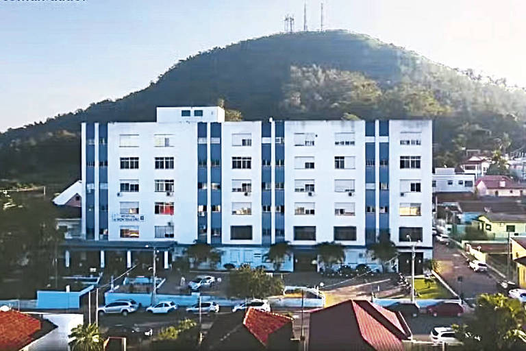 A imagem mostra um edifício de cinco andares， predominantemente branco com detalhes em azul， localizado em uma área urbana. O edifício está cercado por árvores e veículos estacionados. Ao fundo， há uma montanha coberta de vegetação e algumas torres de comunicação visíveis no topo. O céu está claro， indicando um dia ensolarado.